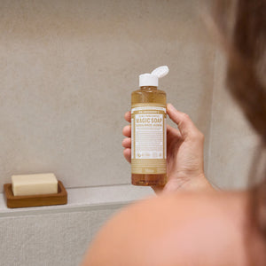 Aroma Jasmim Sândalo da Dr. Bronner's. Mulher a segurar o Sabonete Líquido Biológico e o sabonete barra na prateleira do chuveiro.